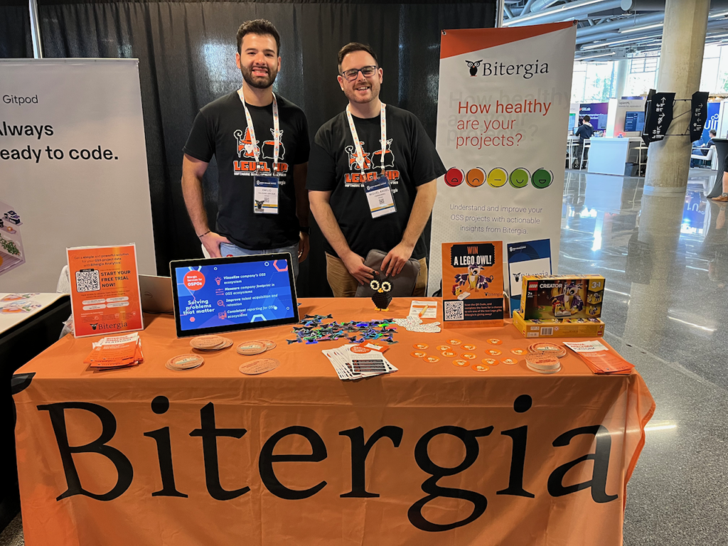 Emilio and Miguel Angel at the Bitergia booth with an orange bitergia tablecloth and table covered in stickers, coasters, and other materials to give away.