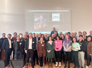 Group picture of the attendees of OFA. Lots of smiling faces with the virtual attendees on the screen in the background.