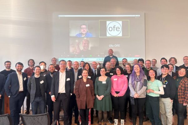 OFA_Group_Picture Group picture of the attendees of OFA. Lots of smiling faces with the virtual attendees on the screen in the background.