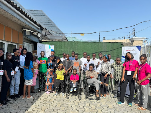 Photograph of a group of people standing, sitting, or in wheelchairs outside in the sunshine.